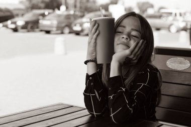 Genç bir kız bir yazlık kafede oturuyor ve meşrubat içiyor.