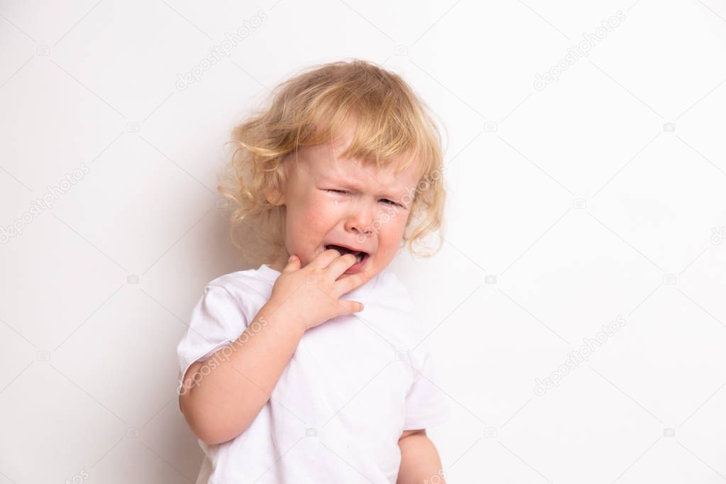 beautiful curly blond baby girl in in white t-shirt crying on a white background. the child has teeth. concept of child care illness. copy space
