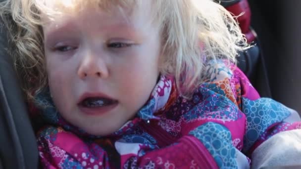 Pequena menina caucasiana com cabelo encaracolado em uma jaqueta quente senta-se em um assento infantil, usando um cinto de segurança andando em um carro chorando em voz alta lágrimas correndo pelas bochechas — Vídeo de Stock