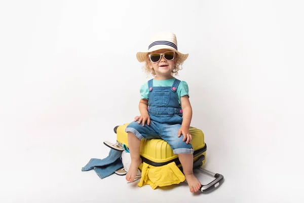 Funny curly little baby girl in a hat, T-shirt and jeans with a yellow suitcase is smiling on white background waiting for an airplane. concept trip with kids. — Stock Photo, Image