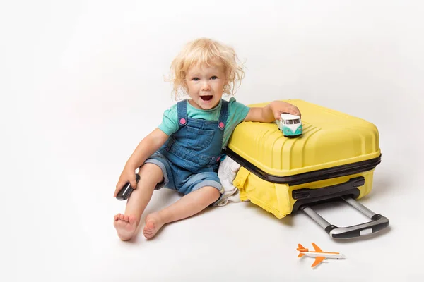 Little Happy kaukaski kręcone dziewczyny w wygodnym denim ubrania dziecko siedzi na żółtym walizce. turysta w koszulce z prostym i samochód w ręku. Pomarańczowa walizka. Wakacje. — Zdjęcie stockowe