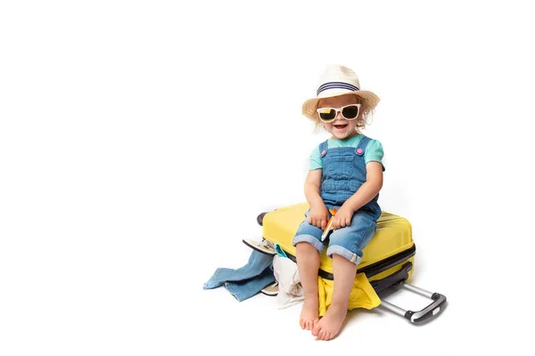 Engraçado encaracolado menina em um chapéu, camiseta e jeans com uma mala amarela está sorrindo no fundo branco esperando por um avião. viagem conceito com crianças . — Fotografia de Stock