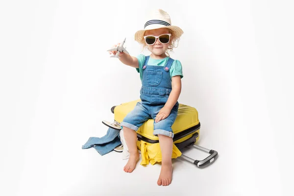 Engraçado encaracolado menina em um chapéu, camiseta e jeans com uma mala amarela está sorrindo no fundo branco esperando por um avião. viagem conceito com crianças . — Fotografia de Stock