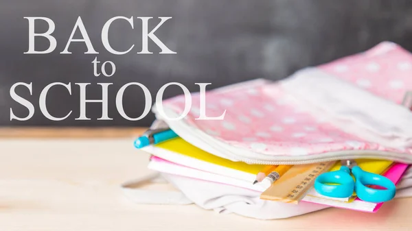 bright open school bag with school supplies lies on a desk against the background of a school blackboard