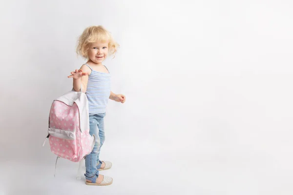Rubio rizado pequeña niña preescolar sostiene una bolsa de color rosa de la escuela sobre un fondo blanco — Foto de Stock