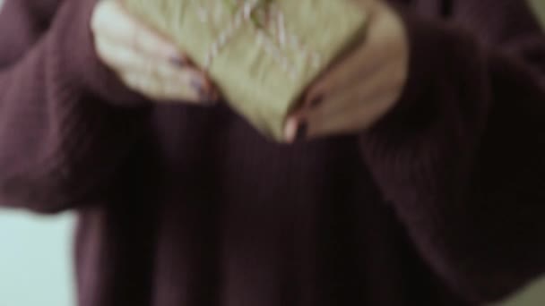 Primer plano de las manos femeninas en un suéter púrpura sosteniendo un pequeño regalo envuelto con papel artesanal. Un pequeño regalo en las manos de una mujer de interior. Profundidad de campo superficial con foco en la cajita . — Vídeo de stock