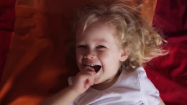 Милая маленькая кудрявая блондинка, смеющаяся в красной постели. Счастливого утра. играя с ребенком — стоковое видео