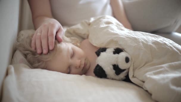 Boldog anya megcsókolja az ágyban fekvő gyermeket és megsimogatja a fejét. — Stock videók