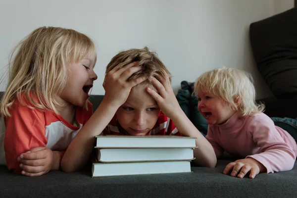 Chico tratar de leer libros, hacer la tarea mientras hermanas gritar hacer ruido —  Fotos de Stock