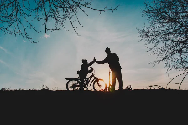 Silhouet van gelukkige vader en zoon paardrijden fiets en scooter bij zonsondergang — Stockfoto