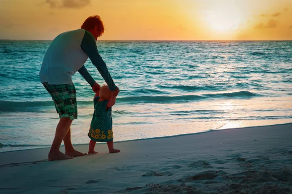 Vader en kleine dochter maken eerste stappen op zonsondergang strand — Stockfoto