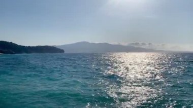 Denizin çarpıcı panoramik görüntüsü. Sudan yansıyan güneş ışığı ufka doğru parıldayan bir yol oluşturuyor. Arka planda, görkemli dağlar berrak bir gökyüzüne yükselir, derinlik ve ihtişam katar.. 