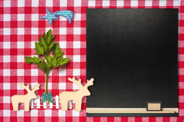 Original background for invitation menu or Christmas recipes. Twig of parsley as a Christmas tree and two wooden reindeer on a kitchen tablecloth and a blank blackboard to write