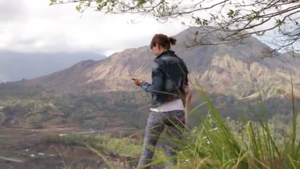 Fiatal nő a smartphone és a hátizsák, egy gyönyörű hegyi háttér előtt. Batur vulkán, Bali. — Stock videók