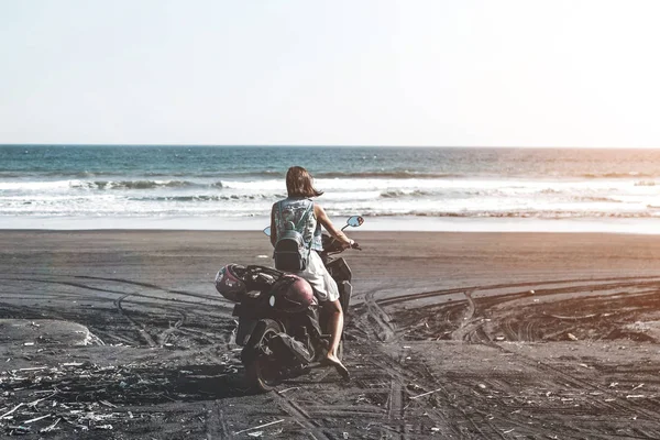 Jovem mulher dirigindo uma scooter na praia com areia preta. Ilha de Bali . — Fotografia de Stock