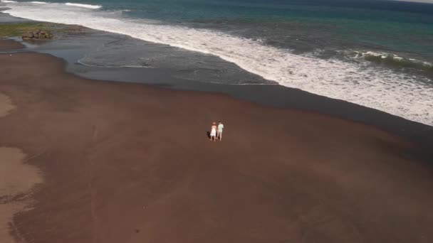 Vídeo de vuelo aéreo de 4K de parejas jóvenes en la playa con arena volcánica negra al atardecer. Isla de Bali . — Vídeo de stock