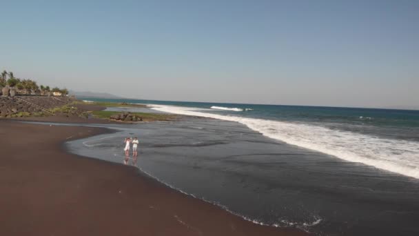 4k antenn flygande video av ungt par använda drönare på stranden med svart vulkansand vid solnedgång. Ön Bali. — Stockvideo