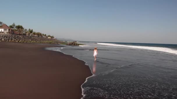 4k repülő légifelvételek fiatal nő fehér ruhában, séta a fekete vulkáni beach, Bali szigetén. — Stock videók