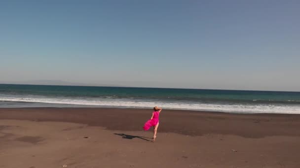 4k εναέρια πλάνα που φέρουν από νεαρή γυναίκα σε ροζ φόρεμα περπάτημα στην μαύρη ηφαιστειακή παραλία, νησί του Μπαλί. — Αρχείο Βίντεο