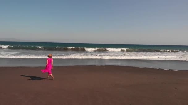 4k luchtfoto vliegende beelden van jonge vrouw in roze jurk lopen op de zwarte vulkanische strand, Bali eiland. — Stockvideo