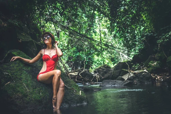 Fiatal nő travele turisztikai piros női fürdőruha, és pózol a dzsungel folyó Szalmakalapot. Bali szigetén. — Stock Fotó