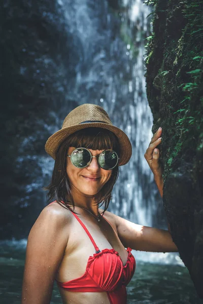 Jonge vrouw toerist in rode zwembroek en stro hoed poseren in de diepe jungle met waterval. Echte avontuur concept. Bali eiland. — Stockfoto