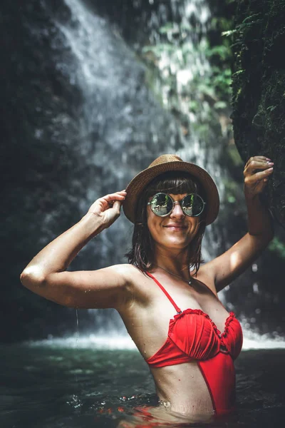 Jonge vrouw toerist in rode zwembroek en stro hoed poseren in de diepe jungle met waterval. Echte avontuur concept. Bali eiland. — Stockfoto