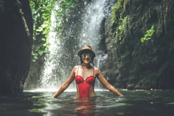 Fiatal nő turisztikai Szalmakalapot és piros fürdőruha-vízesés a mély dzsungelben. Igazi kaland fogalma. Bali szigetén. — Stock Fotó
