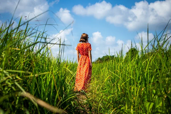 Ung kvinna i röd klänning gå på ett spår med långa gröna gräs. Ön Bali. — Stockfoto