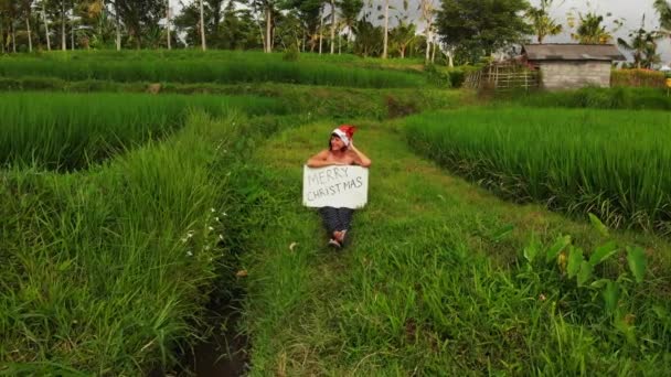 Vídeo de un avión no tripulado volador de una mujer joven con sombrero rojo y pizarra blanca con texto escrito a mano feliz navidad sobre un fondo verde tropical. Saludos de Navidad desde la isla de Bali, Asia, Indonesia . — Vídeo de stock