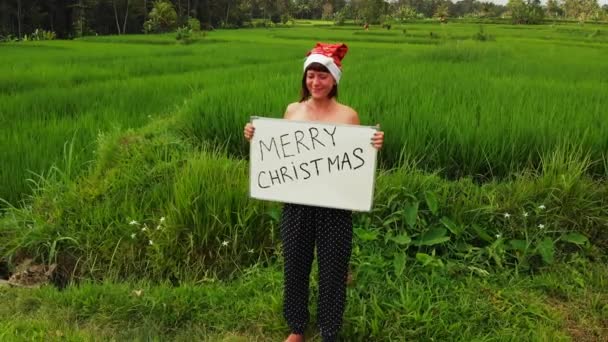 Volar drone video de mujer joven con sombrero rojo y pizarra blanca con texto escrito a mano Feliz Navidad sobre un fondo tropical . — Vídeo de stock