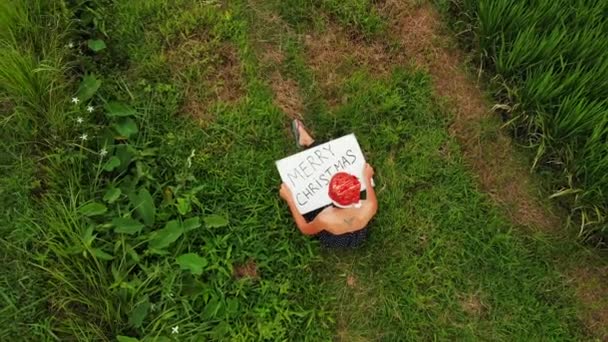 Vidéo drone volant de jeune femme avec chapeau rouge et tableau blanc avec texte écrit à la main joyeux Noël sur un fond tropical .