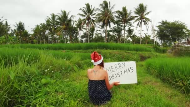 Volar drone video de mujer joven con sombrero rojo y pizarra blanca con texto escrito a mano Feliz Navidad sobre un fondo tropical . — Vídeo de stock