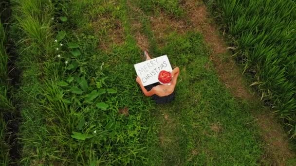 Vidéo drone volant de jeune femme avec chapeau rouge et tableau blanc avec texte écrit à la main joyeux Noël sur un fond tropical .