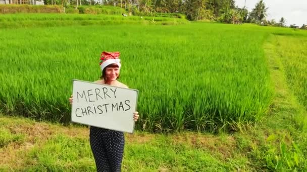 Flying drone video of young woman with red hat and whiteboard with handwritten text merry christmas on a tropical background. — Stock Video