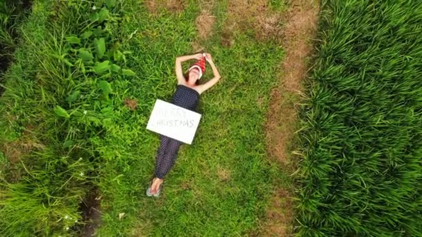 Vídeo de un avión no tripulado volador de una mujer joven con sombrero rojo y pizarra blanca con texto escrito a mano feliz navidad sobre un fondo verde tropical. Saludos de Navidad desde la isla de Bali, Asia, Indonesia . — Vídeos de Stock