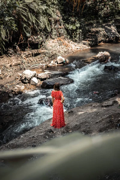 Mujer joven en vestido rojo largo posando en un río de montaña en lo profundo de la selva tropical de la isla de Bali, Indonesia . — Foto de Stock