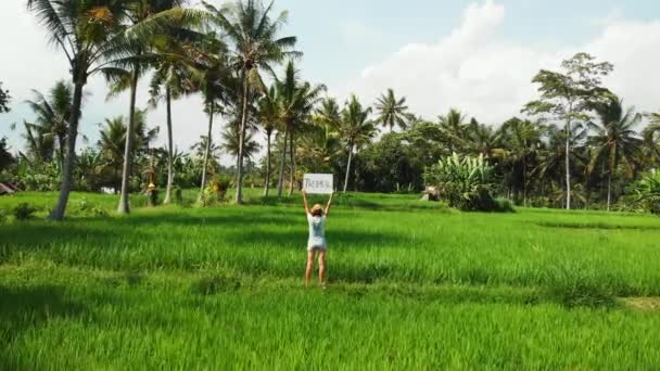 Young woman outdoors with whiteboard and handwriting word tropical on it. Drone flying footage. Green bright tropical background. Beautiful tropical travel concept. Bali island. — Stock Video