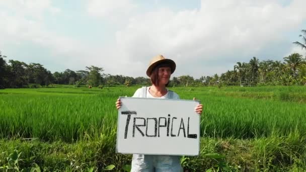 Jovem mulher ao ar livre com quadro branco e caligrafia palavra tropical sobre ele. Imagens de drones a voar. Fundo tropical verde brilhante. Belo conceito de viagem tropical. Ilha de Bali . — Vídeo de Stock