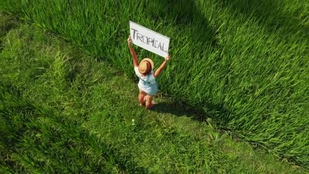 Young woman outdoors with whiteboard and handwriting word tropical on it. Drone flying footage. Green bright tropical background. Beautiful tropical travel concept. Bali island. — Stock Video