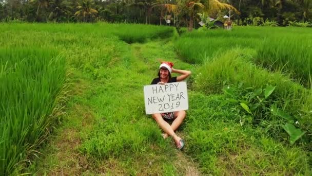 Frohes neues Jahr 2019. 4k fliegendes Drohnenvideo einer jungen Frau mit Weihnachtsmütze und Whiteboard mit handgeschriebenem Text, die inmitten eines grünen Reisfeldes auf der Insel Bali posiert. — Stockvideo