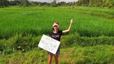 Mutlu yeni yıl 2019. Noel Baba şapkası olan kadın ve beyaz tahta el yazısı metin yeşil pirinç alan Bali Adası arasında poz ile video 4k uçan robot.