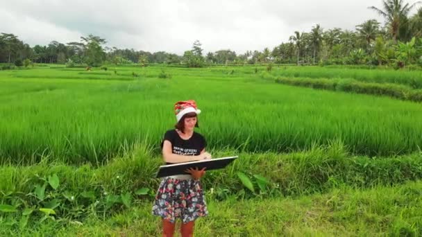 Gelukkig Nieuwjaar 2019. 4k vliegende drone video van jonge vrouw met kerstmuts en whiteboard met handgeschreven tekst poseren onder groene rijst veld op Bali eiland. — Stockvideo