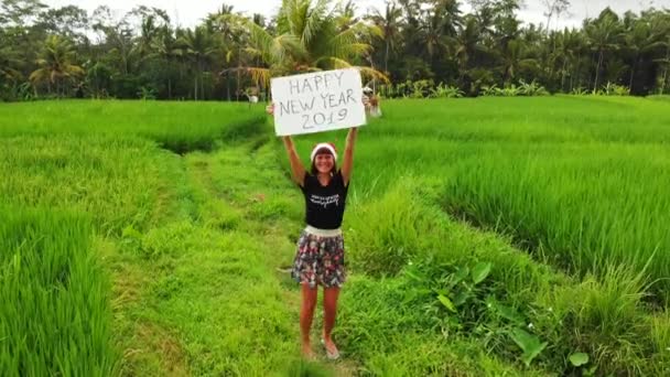 Boldog új évet 2019. 4k repülő drone videó a fiatal nő, santa kalap és a faliújság, a kézzel írott szöveg jelentő között zöld rizs mező Bali szigetén. — Stock videók