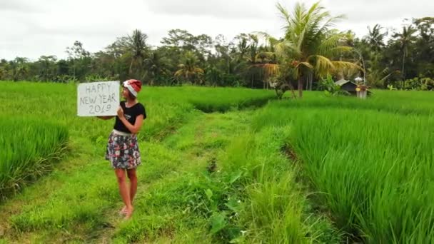Feliz Año Nuevo 2019. 4K vuelo drone video de mujer joven con sombrero de santa y pizarra blanca con texto escrito a mano posando entre el campo de arroz verde en la isla de Bali . — Vídeos de Stock