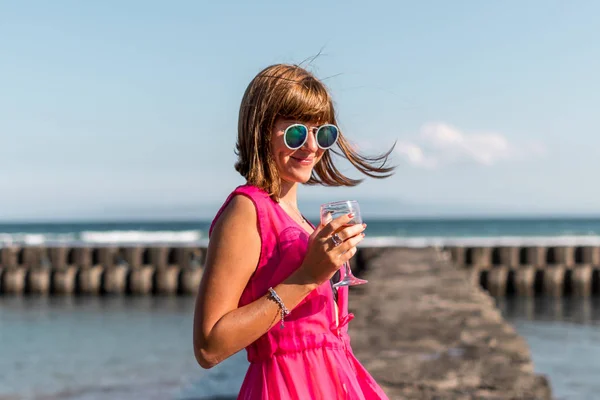 Молодая женщина в длинном красном платье и с бокалом вина позирует на морском фоне . — стоковое фото
