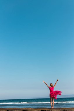 Genç kadın uzun kırmızı bir elbise ve bir kadeh şarap deniz arka plan üzerinde poz ile.