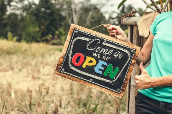 Open sign board in woman hands. Bali island background.