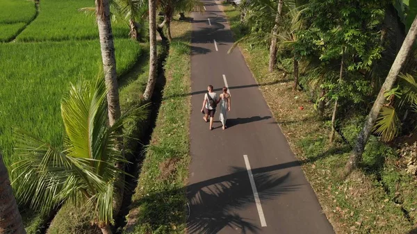 Vista aérea de una joven pareja de turistas caminando por la carretera entre palmeras de coco. Isla de Bali . — Foto de Stock