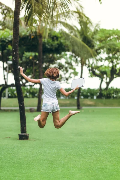 Young freelancer woman jumping with laptop in the beautiful tropical green park of Bali island.
